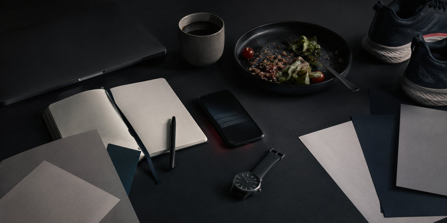 A dark, minimal desk with a notebook, coffee, a simple meal, running shoes and a watch